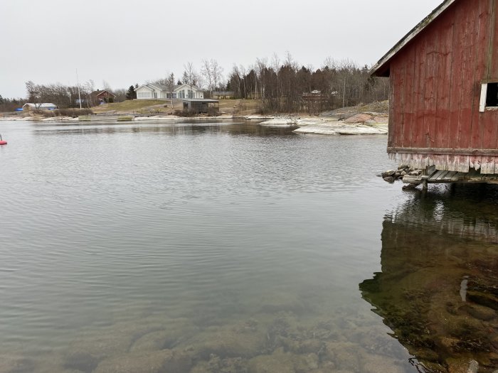 Ett rödfärgat sjöbod vid vattnet, med genomskinlig sjö och hus i bakgrunden under en mulen himmel.