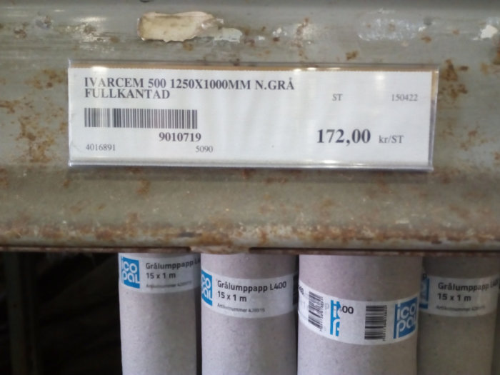 Etikett för IVARCEM fibercementprodukter med prisinformation på en hylla ovanför byggmaterialrullar.