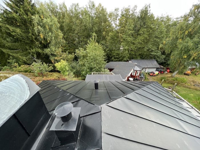Utsikt över ett nylagt svart plåttak med ventilation och grönskande trädgård i bakgrunden.