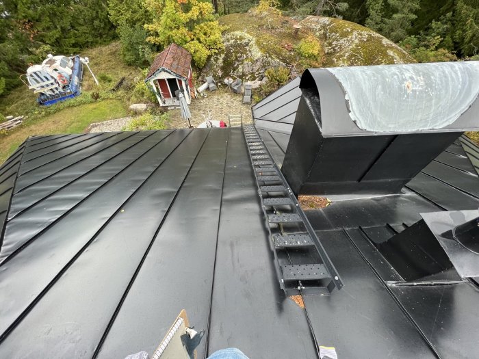 Nyinstallerat svart plåttak med stege och skorsten, trädgård i bakgrunden