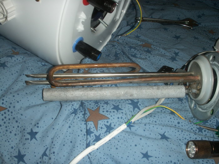 Ljusgrå tjock cylinder som är en offeranod framför rostiga värmeelement på ett blåstjärnigt tyg.