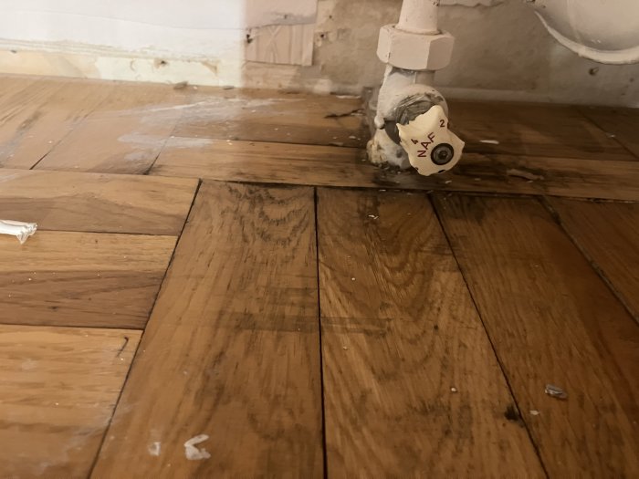 Skadat trägolv med vattenskada kring vitmålad radiatorventil.