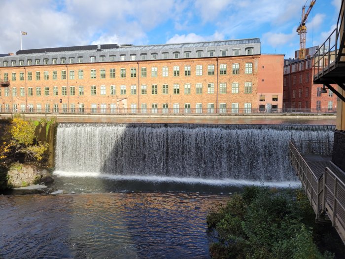 Vattenfall framför gammal tegelbyggnad, med en kran och del av bro synliga.