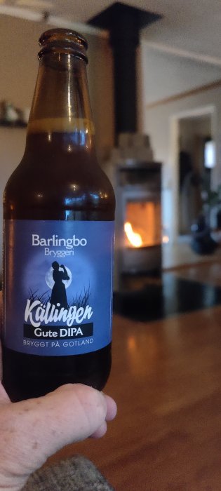 En hand håller en ölflaska med etiketten "Källingen Gute DIPA" med en brasa i bakgrunden.