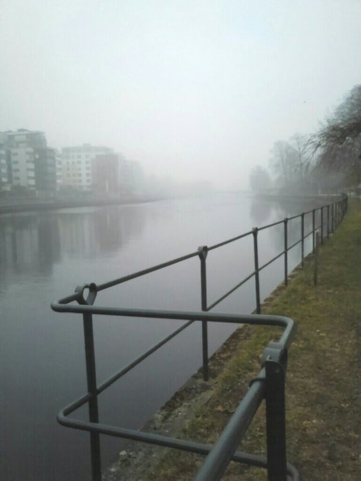 Dimmig vy längs en flod med svart metallräcke i förgrunden och konturer av byggnader i bakgrunden.