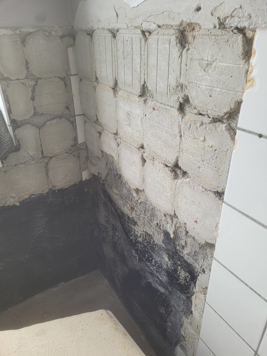 Hörn av ett badrum under renovering med oputsade betongblock och vattentätning på väggarna.
