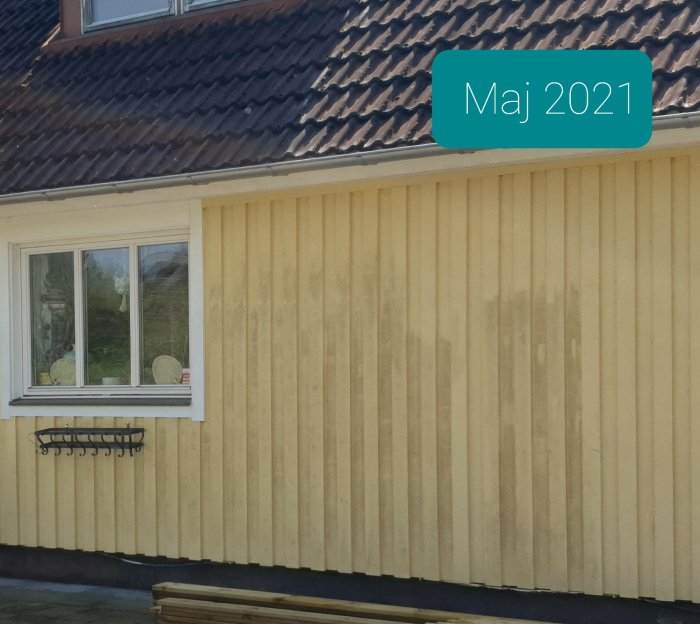 Husfasad med träpanel och ett fönster, texten "Maj 2021" i övre högra hörnet.