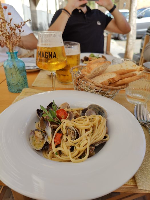 En tallrik spaghetti vongole med musslor och tomater, ölglas på bordet och en person i bakgrunden.