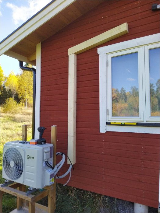 Rödmålad stugvägg med nyinstallerade träsnickerier runt ett fönster och en värmepump framför, omgiven av natur.