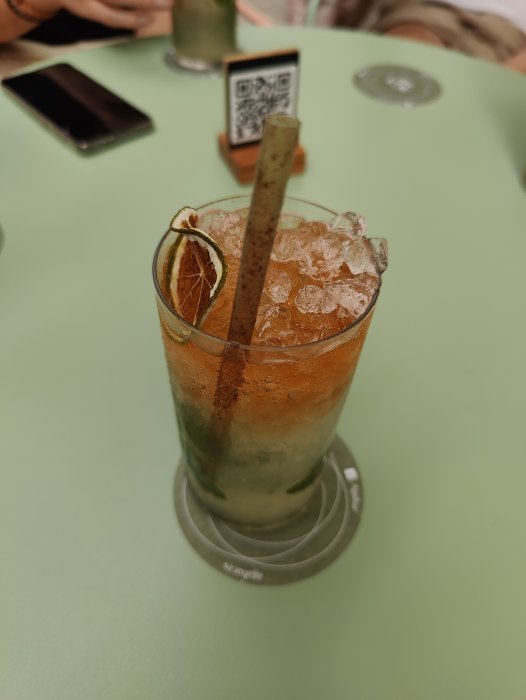 Ett glas mojito med is, lime och sugrör på ett grönt bord.