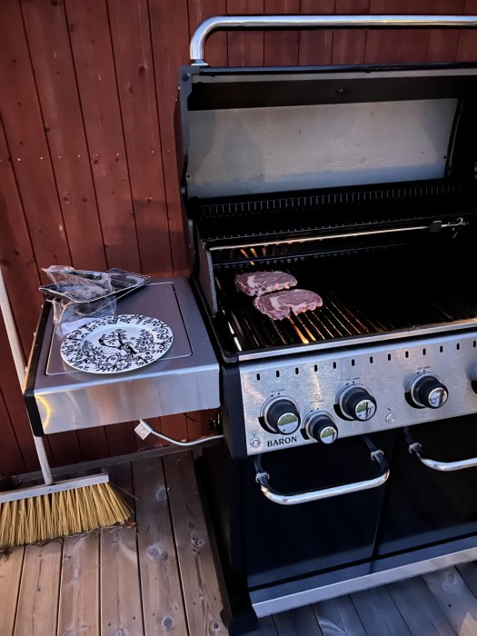 Gasolgrill med stekta biffar på gallret, stängd överhylla och ett dekorativt tallrik på sidobordet.