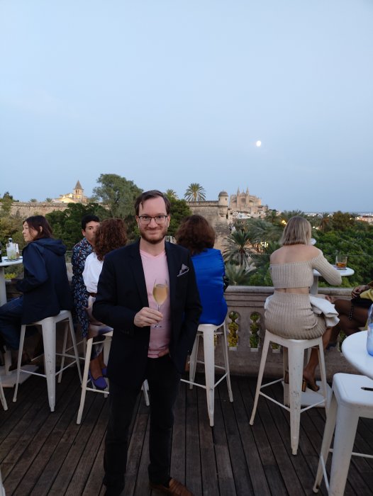 Man i kostym håller ett glas cava på en terrass med utsikt över en historisk byggnad och månen.