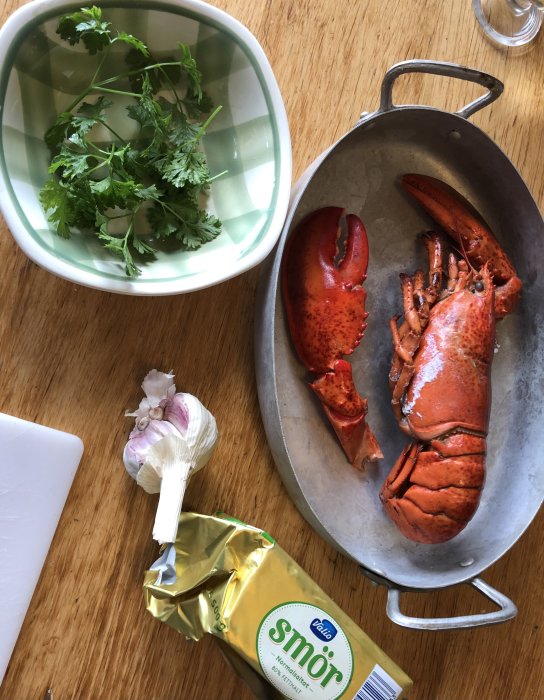 Hummer på serveringsfat bredvid vitlöksklyftor, färsk persilja och ett paket smör på träbord.