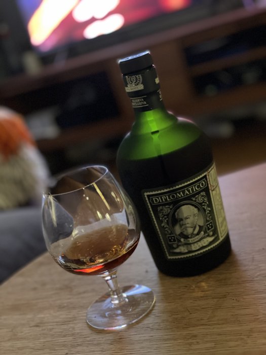 Flaska av Diplomatico rom bredvid ett cognacsglas på ett bord med en suddig TV i bakgrunden.