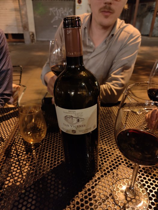 Flaska Rioja-vin med etikett "San Vicente" på bord med glas, person i bakgrunden.