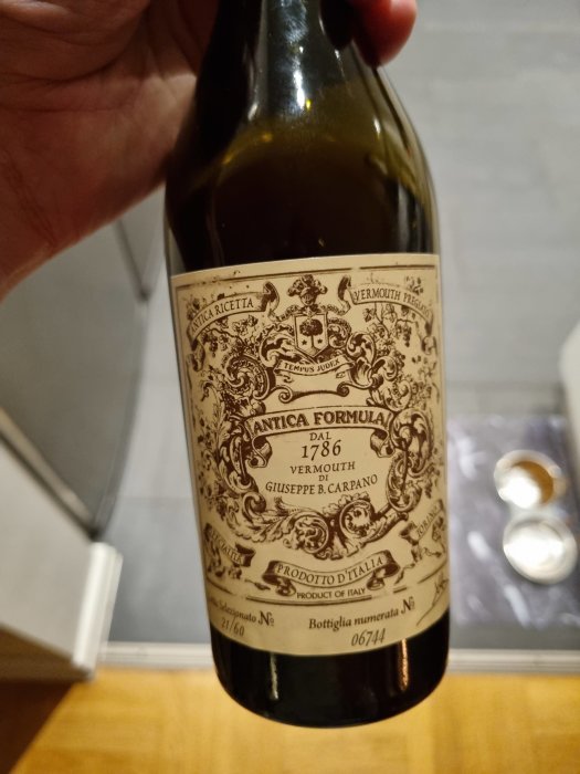 Flaska med Antica Formula vermouth hållen i handen framför ett köksskåp.