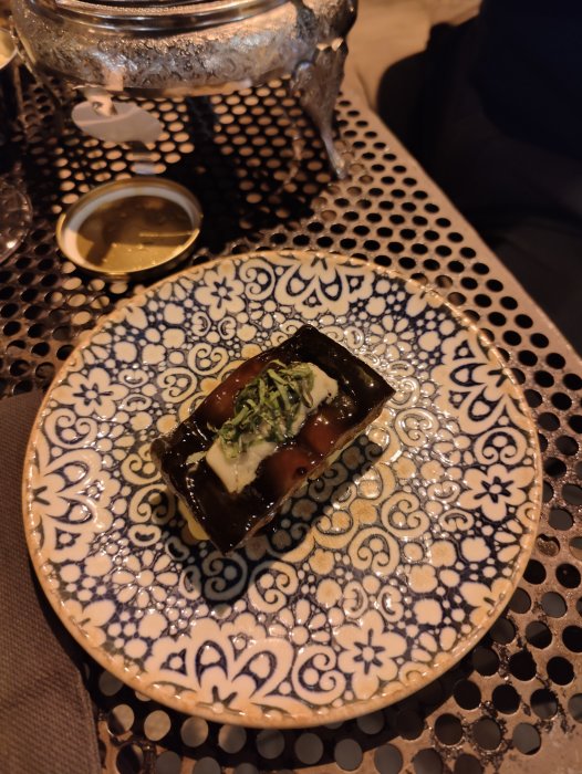 Ål och anklever på dekorerad tallrik med mönster, serverad på ett metallställ på bord.