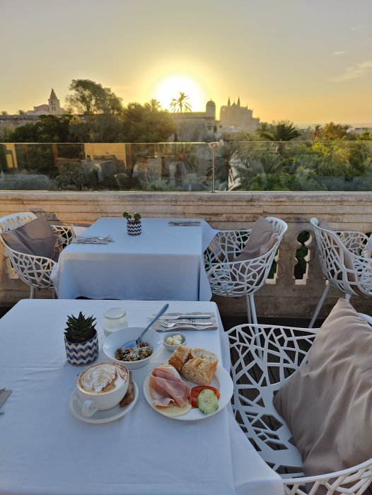 Hotellfrukost på terrass med utsikt över palmträd, kyrktorn och soluppgång.