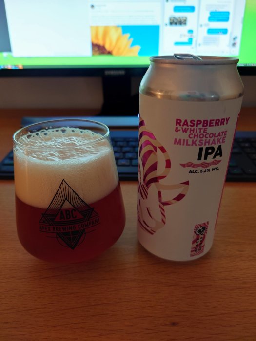 Ölglas med skum öl bredvid en burk Raspberry & White Chocolate Milkshake IPA på ett skrivbord.