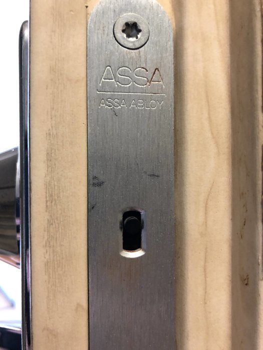 Närbild av ett befintligt ASSA ABLOY dörrlås monterat i en träkarm, med översikt av låskista och nyckelhål.