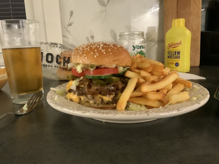 Juicy hamburger with cheese, toppings, and fries on a plate, with mustard and a glass of beer in the background.