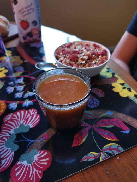 Glas med äpple/morotsjuice bredvid en skål granola på färgglatt blommönstrat bord.