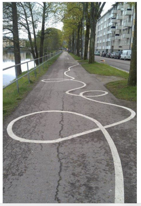 Gångväg vid vatten med oregelbundet kritvit målningsmönster på asfalten liknande konst.