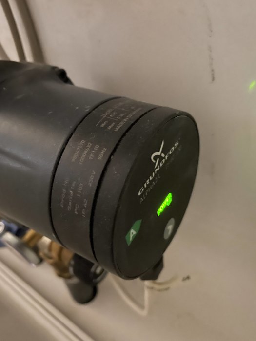 Svart cirkulär vattenpump vid vägg med tänd grön strömindikatorlampa.