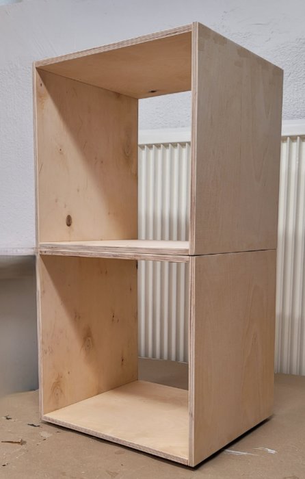 Nytillverkad, stapelbar vedkorg i plywood, utan handtag, står framför en radiator.