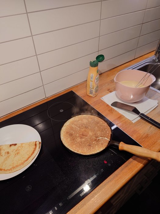 Stekpanna med pannkaka på spis, ingredienser och en bunke med smet på en köksbänk.