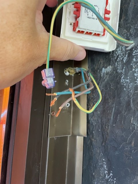 Hand håller elektriska kablar framför en öppen vägguttag med synliga ledningar och anslutningspunkter.