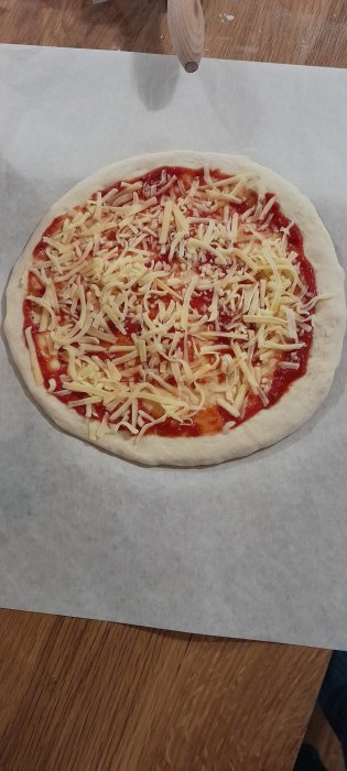 Ostbelagd, tomatsåsbaserad pizzadeg på bakplåtspapper bredvid en kavel.