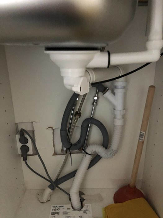 Rörinstallation under diskho med anslutningar, sladdar och plungers, behov av städning och organisation.