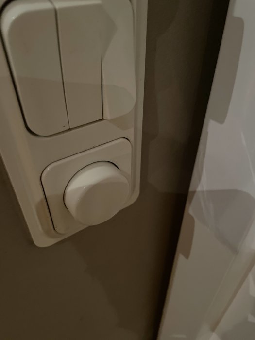 Vit termostatinställningsknapp på en vägg intill ljusströmbrytare.