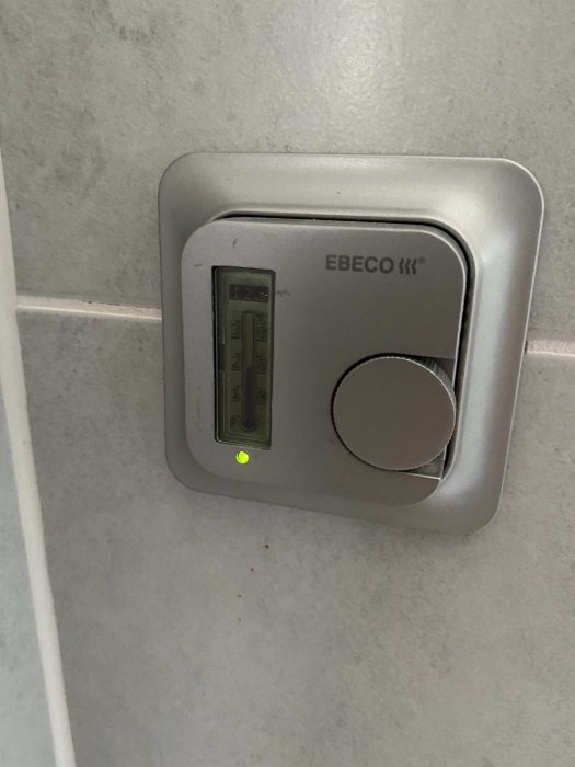 EBECO termostat för golvuppvärmning monterad på en grå vägg med en lysande grön indikatorlampa.