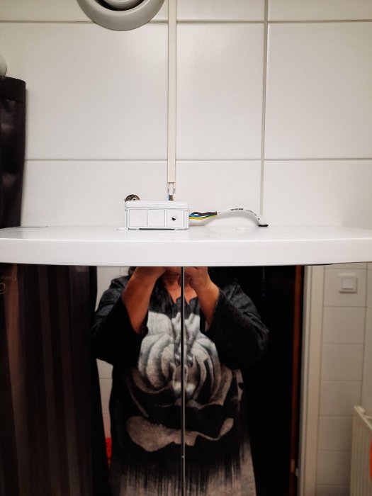 Eluttag monterat på kakelvägg med öppna kablar, spegelreflektion av person fotograferande.