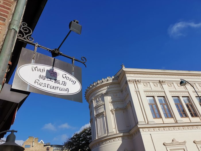 Skylt för Mui Gong Restaurang med kinesiska tecken mot en klarblå himmel och klassisk byggnadsfasad.