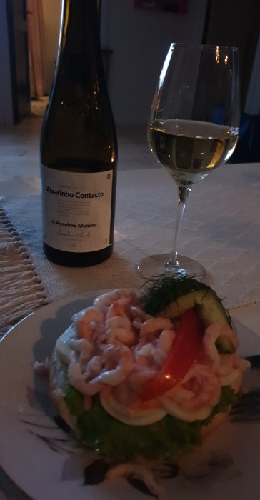 Räksmörgås med sallad och tomat på tallrik bredvid en flaska vitvin och ett glas.