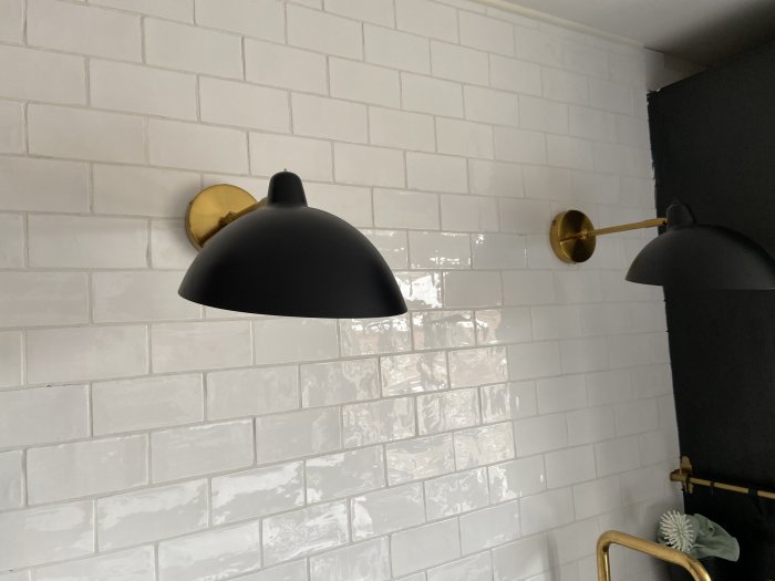 Svart vägglampa med guldfärgad armatur på en vit tegelvägg.