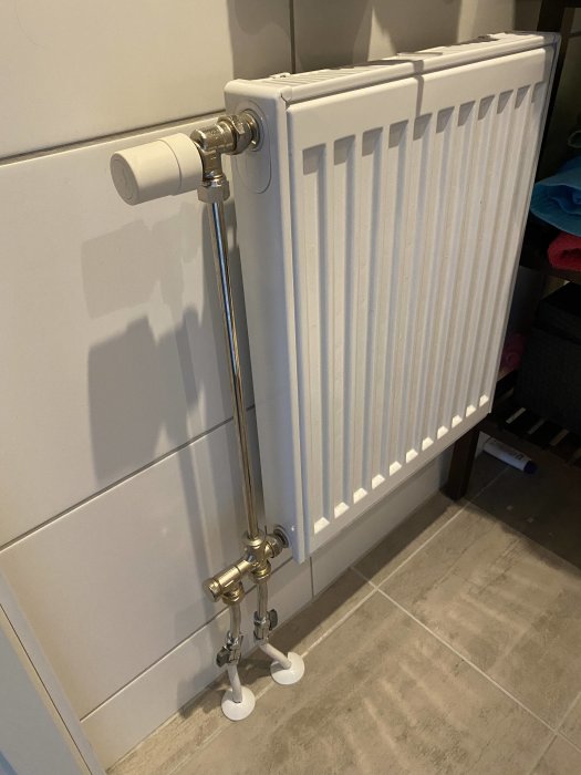 Vit elementradiator i ett rum med grått kakelgolv och anslutna kromade rör.