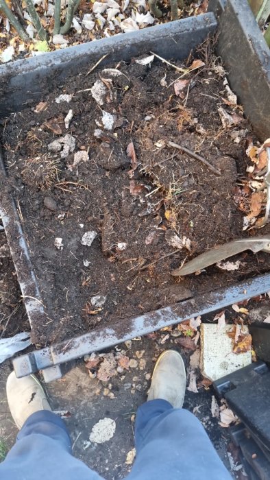 Fötter i arbetsstövlar står framför en kompostlåda med jord och löv, som syns i förgrunden.