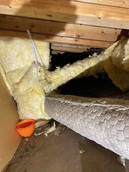 Grått ventilationssrör går genom isolerad vind, orange plastburk på golvet.