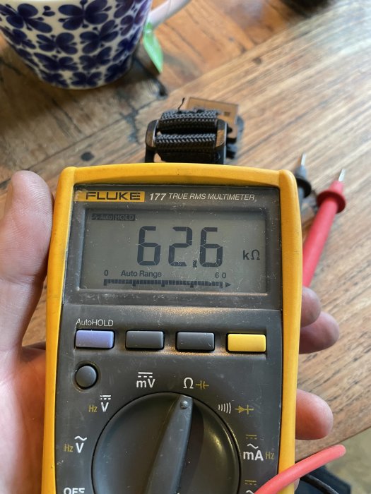 En hand håller en Fluke 177 multimeter som visar ett motstånd på 62,6 kOhm, vid sidan av en kopp och avtagbara motorkol.