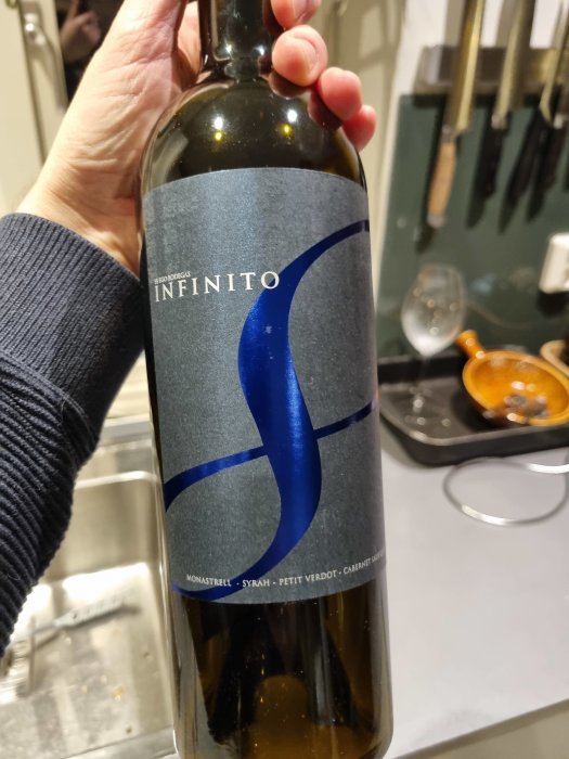 Hand håller en flaska vin med etiketten "INFINITO", druvsorterna Monastrell, Syrah, Petit Verdot och Cabernet Sauvignon listade.
