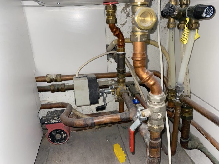 Rörinstallation under varmvattenberedare med diverse kopplingar, ventiler och en röd box.