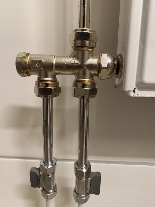 Två kromade rör kopplade till en mässingsfördelare monterad på en vägg, möjlig ful-lösning för elgolvvärme.