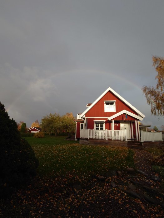 Röd trävilla med vita knutar under en himmel med regnbåge, omgivet av en höstträdgård med löv.