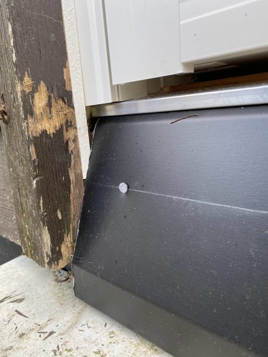 Närbild på ett svart fönsterbleck av plåt med synlig skarv och skruv mot sliten träkarm och vit fönsterram.