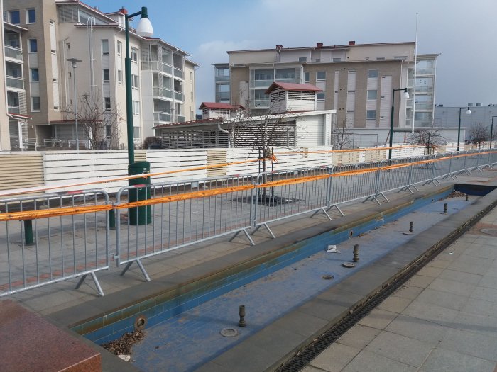 Konstruktionsområde med orangea staket nära en tömd, blå pool med stadshus i bakgrunden.