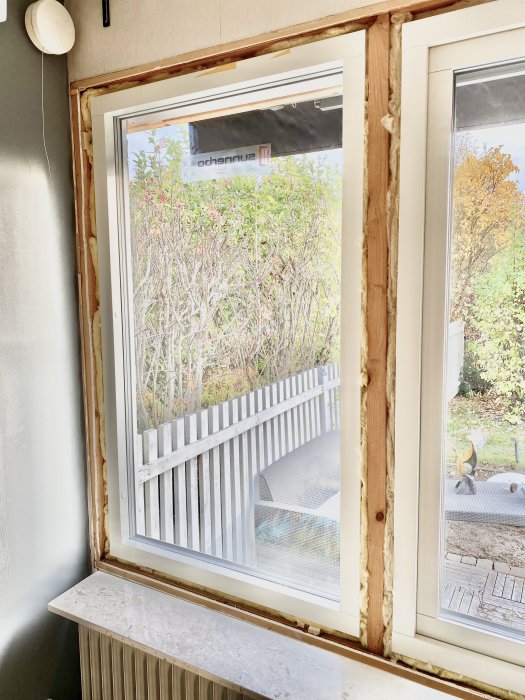 Öppet vardagsrumsfönster med synlig träram och utsikt över trädgård, belyst för att visa höjdskillnad jämfört med intilliggande fönster.